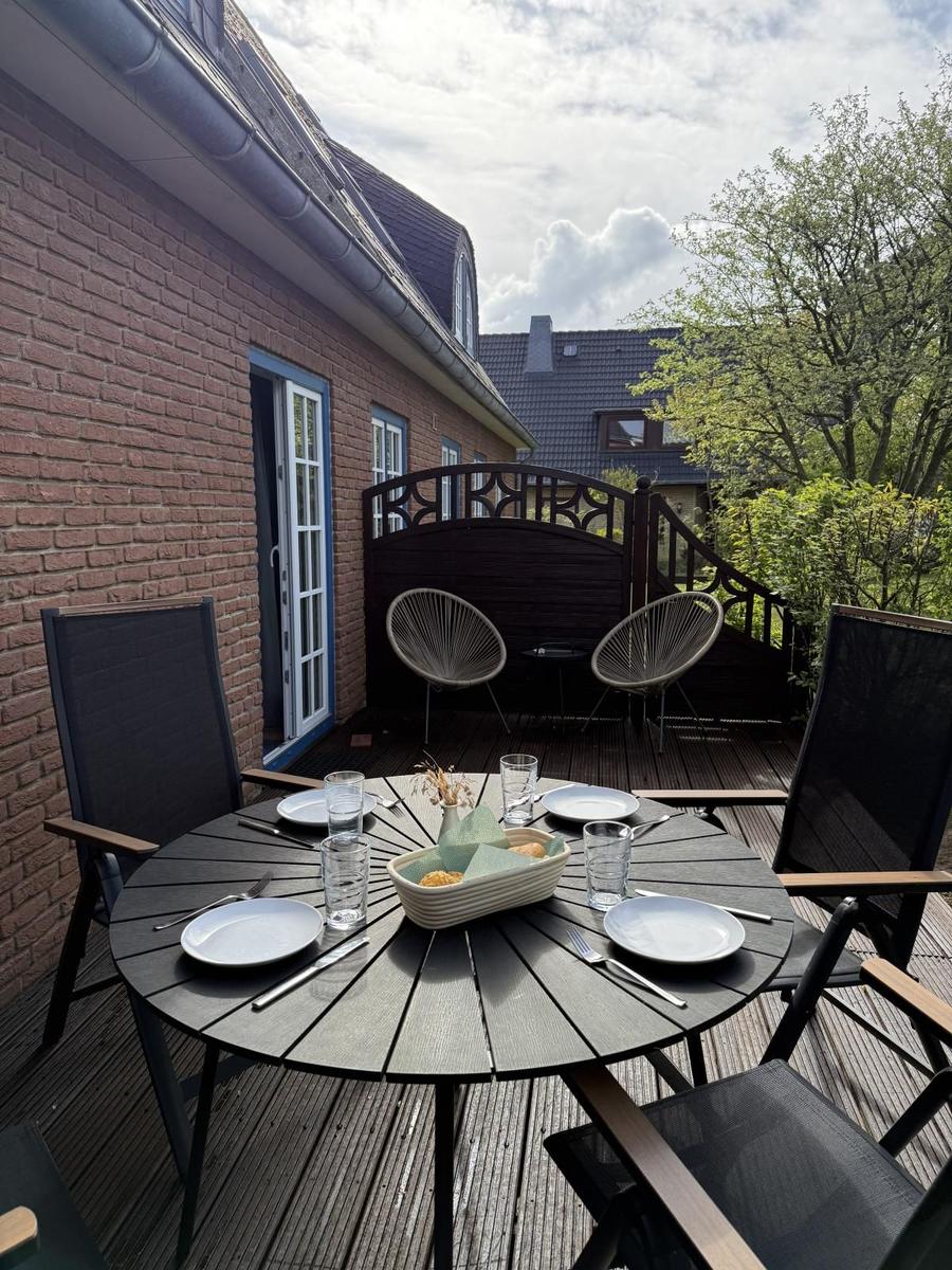 Rundtisch mit Stühlen auf Holzterrasse neben Backsteinhaus mit Garten.
