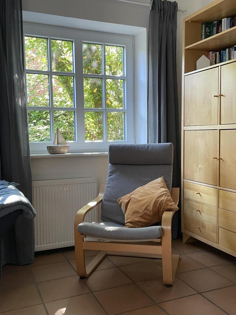 Ein Raum mit Sessel, Fenster und Holzschrank. Draußen sind Bäume sichtbar.