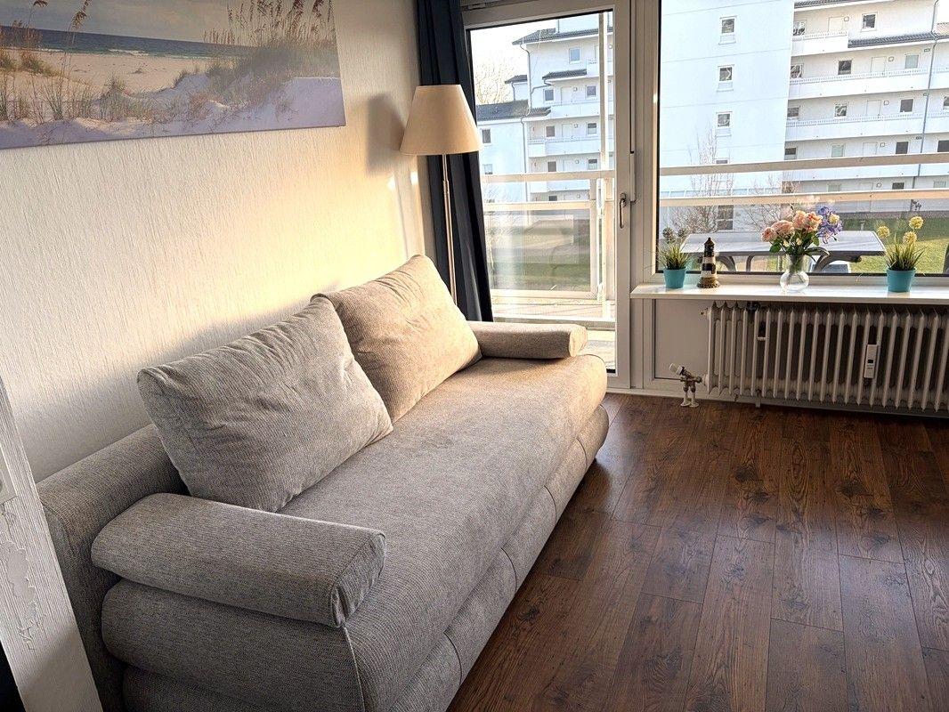 Ein grauer Sofa in einem Raum mit Holzboden und großem Fenster. Auf dem Fensterbrett stehen Pflanzen.