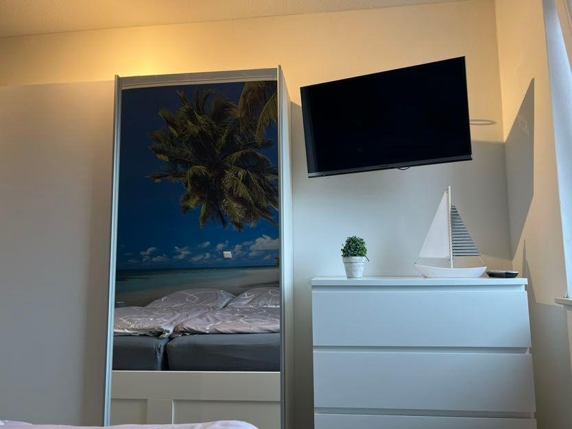 Schlafzimmer mit Spiegeltür, Fernseher und Schrank. Auf Schrank steht Segelbootmodell und Pflanze.