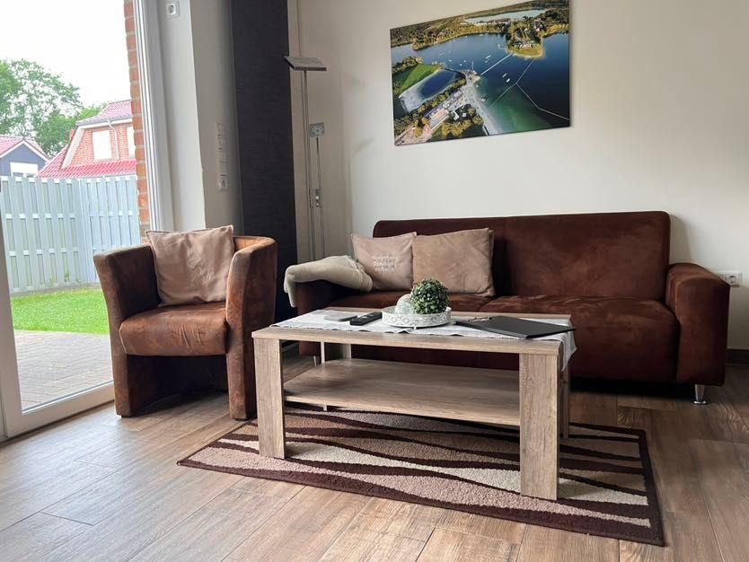 Wohnzimmer mit braunem Sofa, Sessel, Couchtisch und Wandbild. Durchgang zur Terrasse.