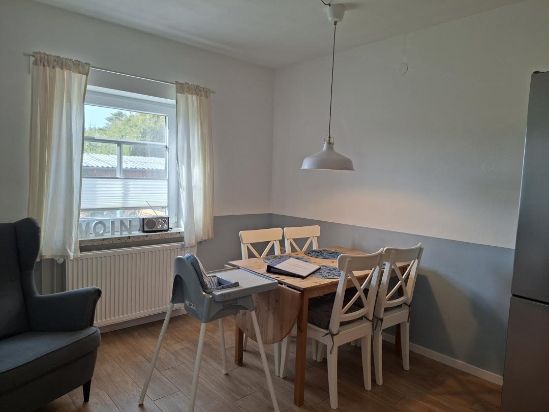Essbereich mit Holztisch, Stühlen und Sessel. Fenster mit Vorhängen und Küchenzeile.