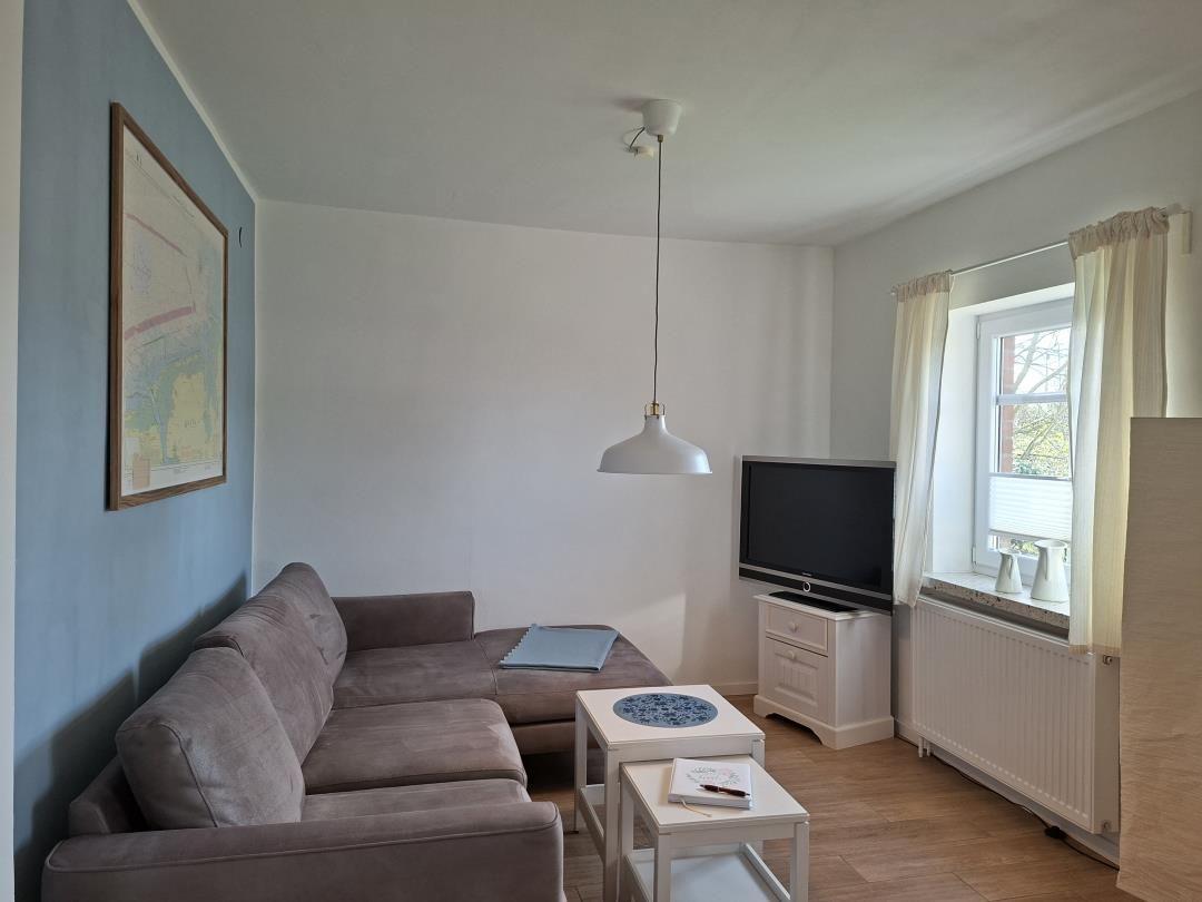 Wohnzimmer mit Sofa, Fernseher und Tischen. Fenster mit Vorhängen und Wandbild.