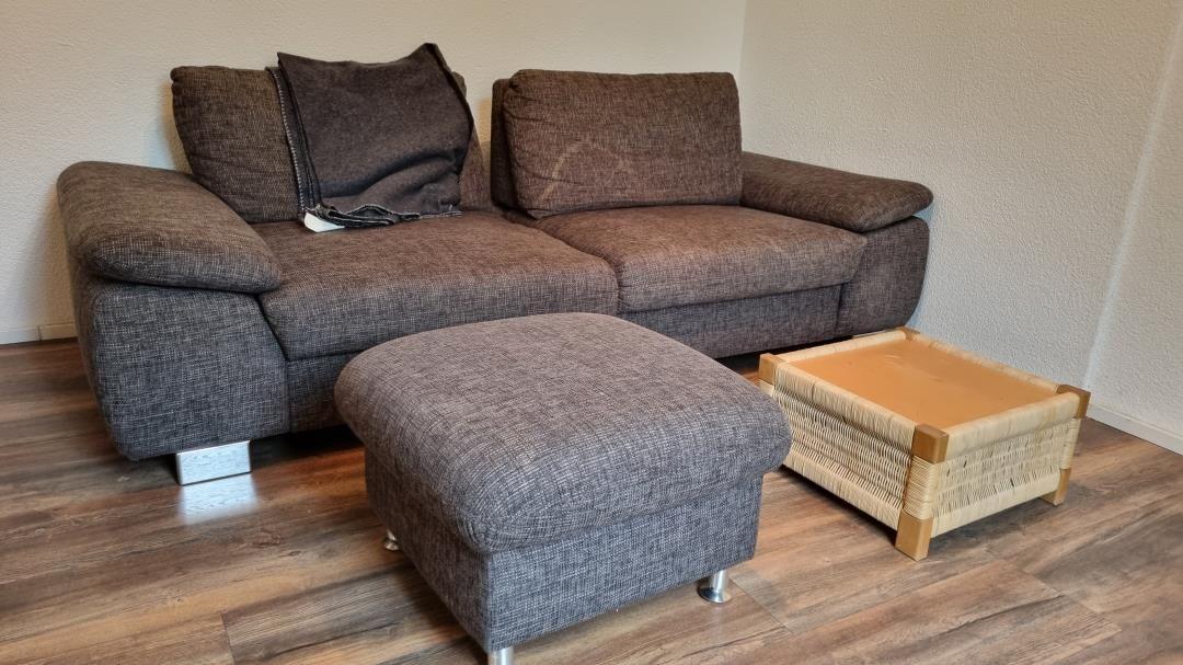 A brown sofa with cushions, an ottoman, and a wicker table on wooden floor.