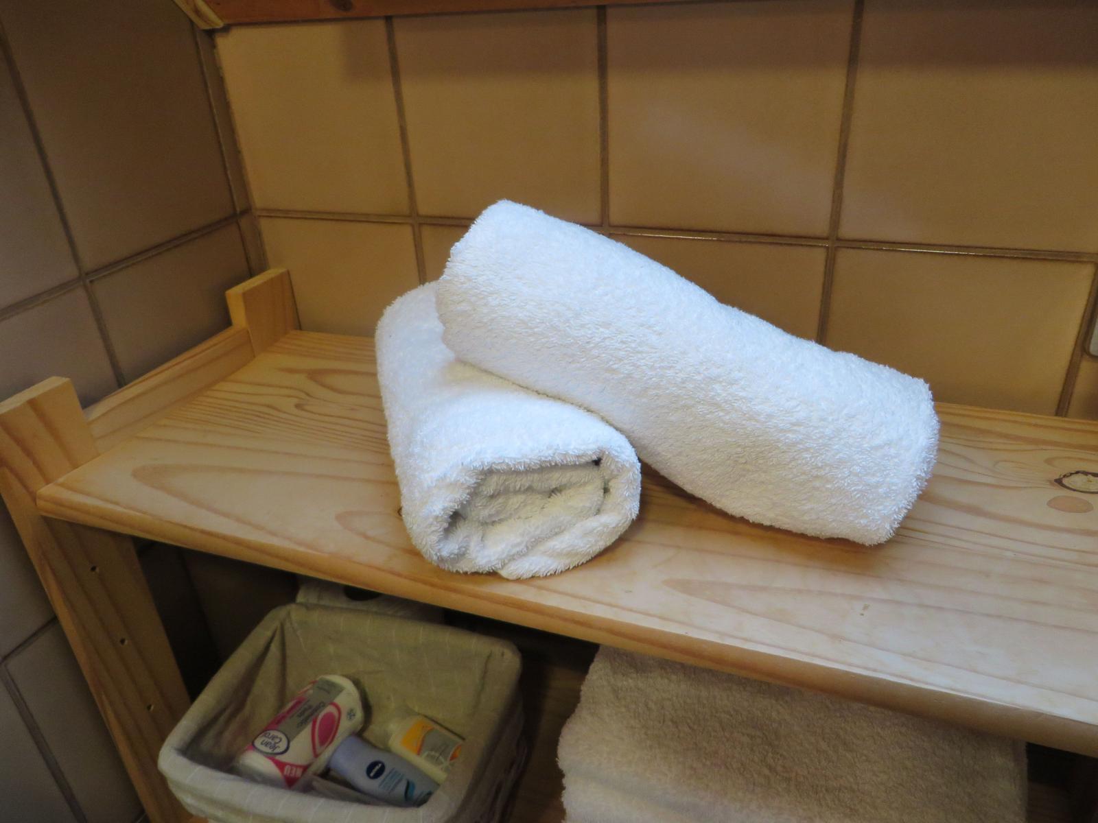Zwei weiße Handtücher liegen auf einem Holzregal. Unter dem Regal befindet sich eine Schachtel mit Pflegeprodukten.