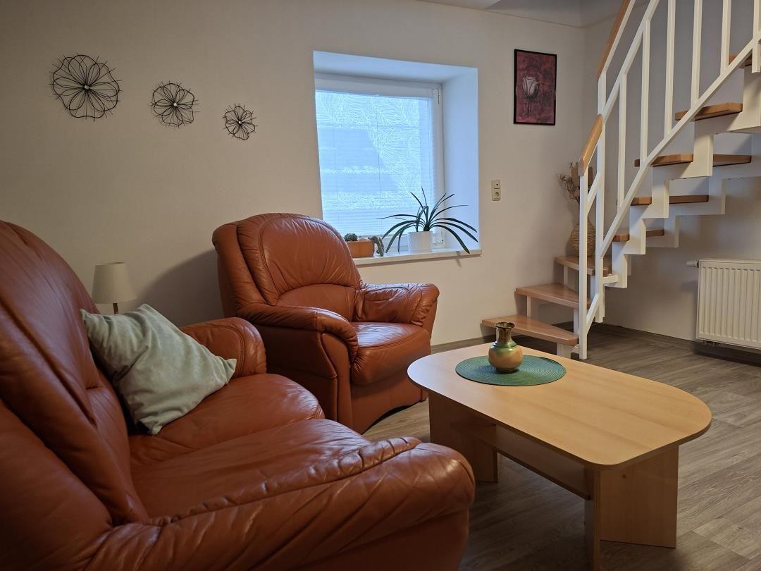 Wohnzimmer mit braunem Ledersofa, Sessel, Tisch und Treppenhaus.
