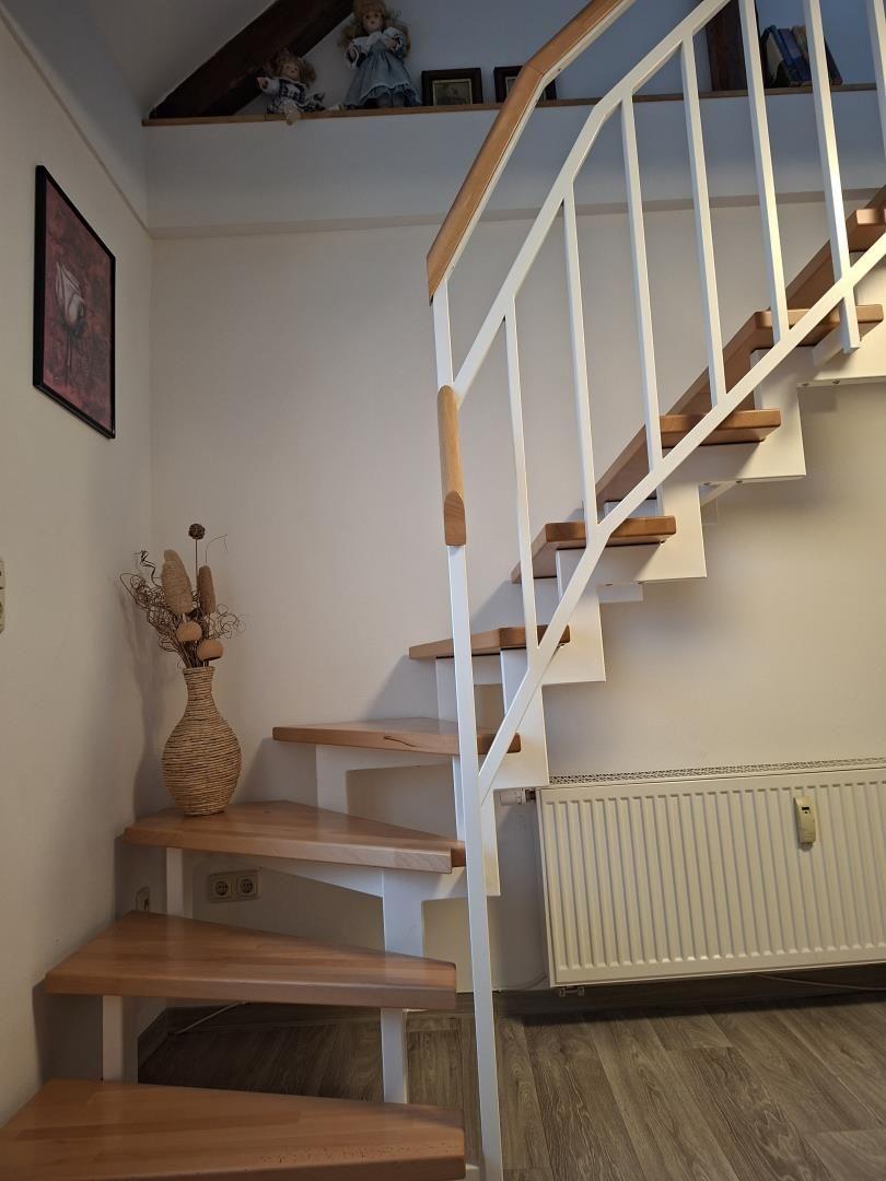 Treppenhaus mit Holzstufen und weißer Metallbrüstung. An der Wand hängt ein Bild und ein Vasenarrangement.