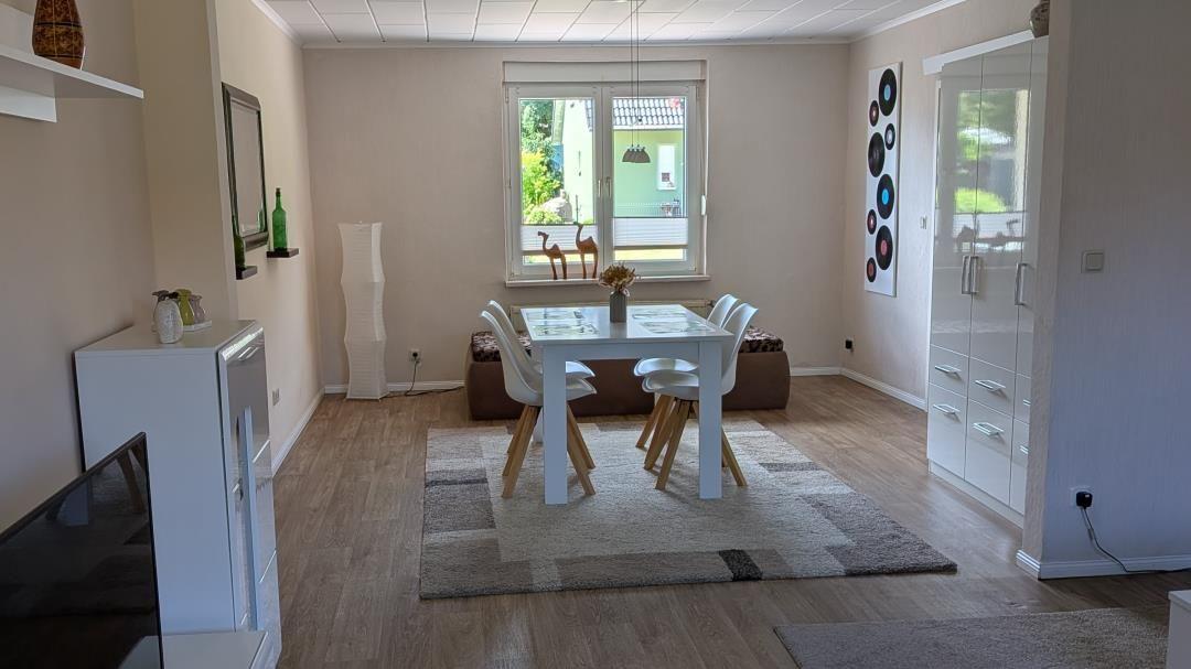 Essbereich mit Tisch, Stühlen und Fenster. Hintergrund: Wohnzimmer mit Fernseher und Schränken.