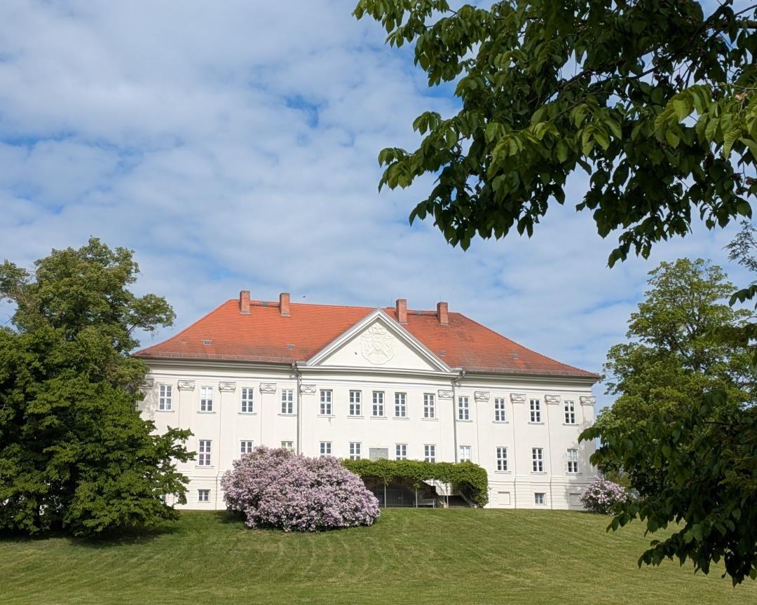 Weißes Gebäude mit rotem Dach, umgeben von Gras und Bäumen mit blühenden Sträuchern.