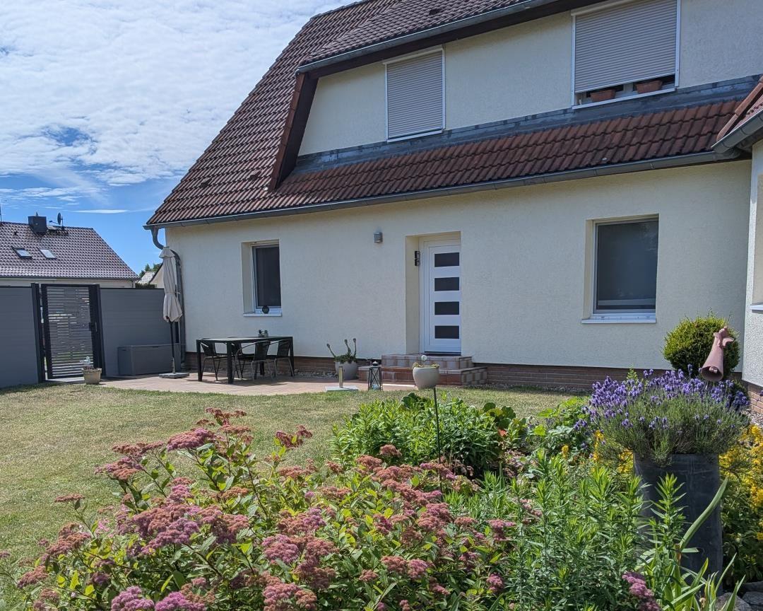 Haus mit Garten, Terrasse und Sitzgelegenheit. Blumenbeete und Pflanzen im Vordergrund.
