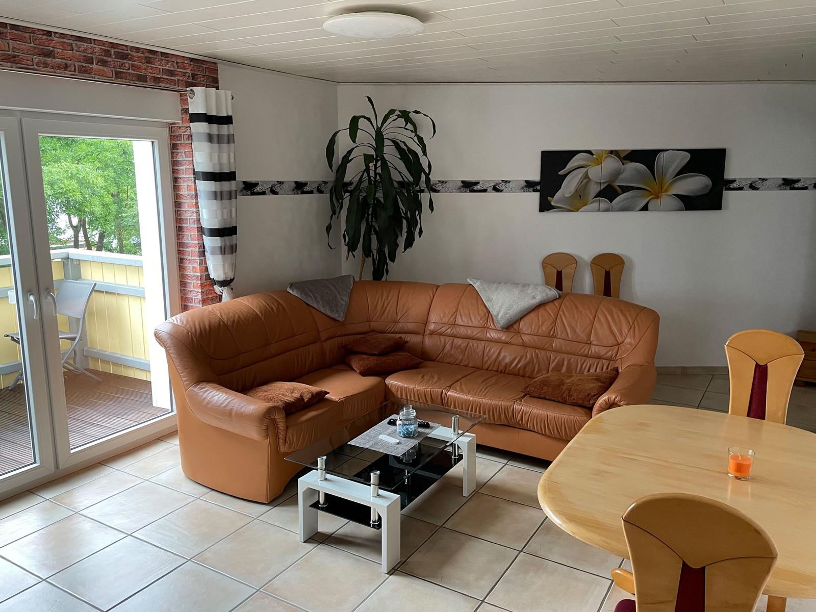 Living and dining area with brown sofa, glass table, and wooden table. Entrance to terrace.