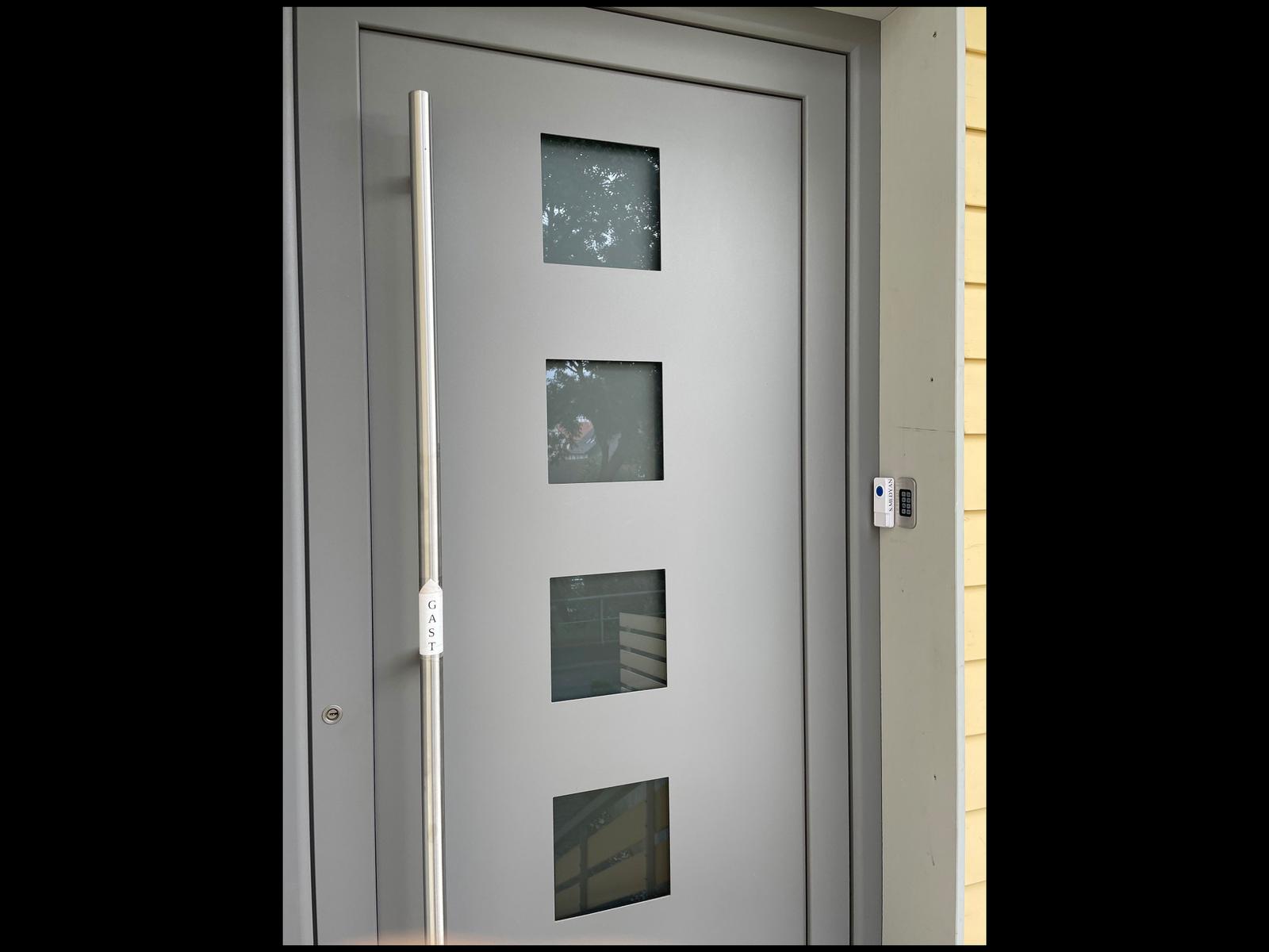 Modern gray front door with four glass panels and handle. A keypad lock is mounted beside the door.