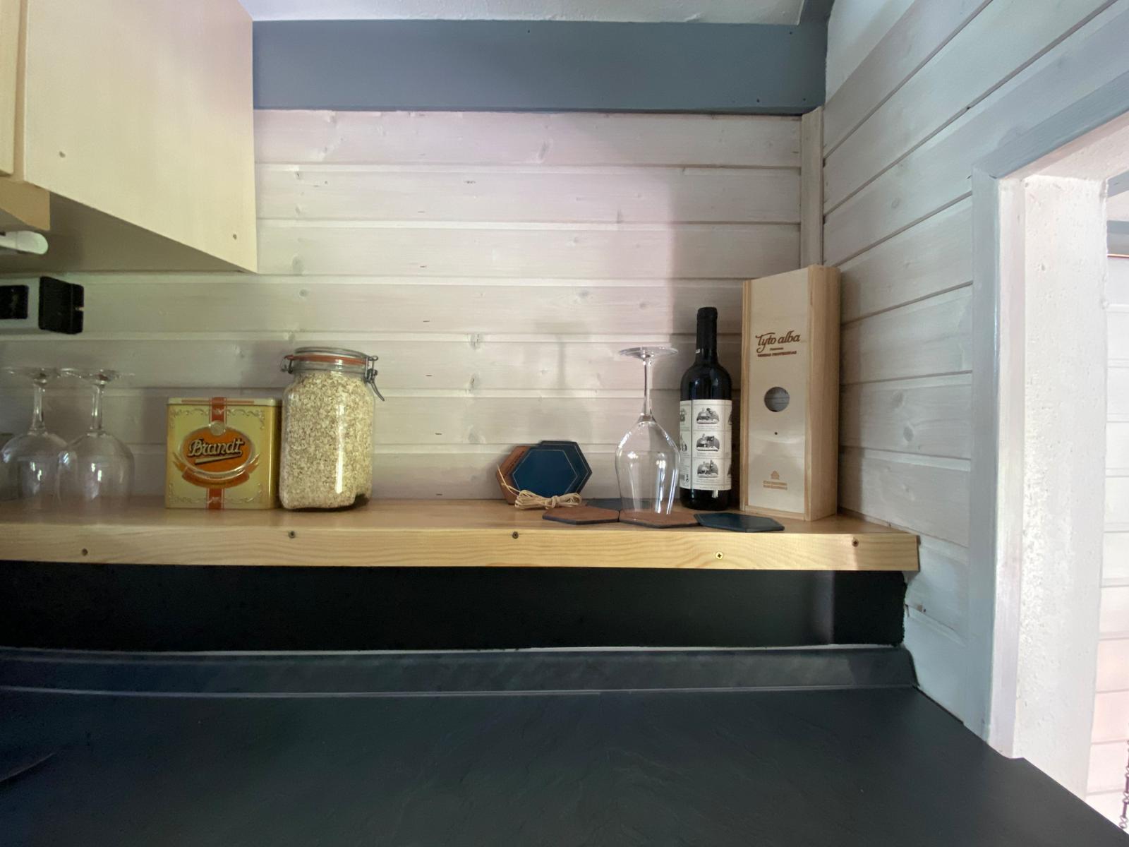 Holzregal mit Glasflasche, Weinflasche und Küchenutensilien an der Wand.