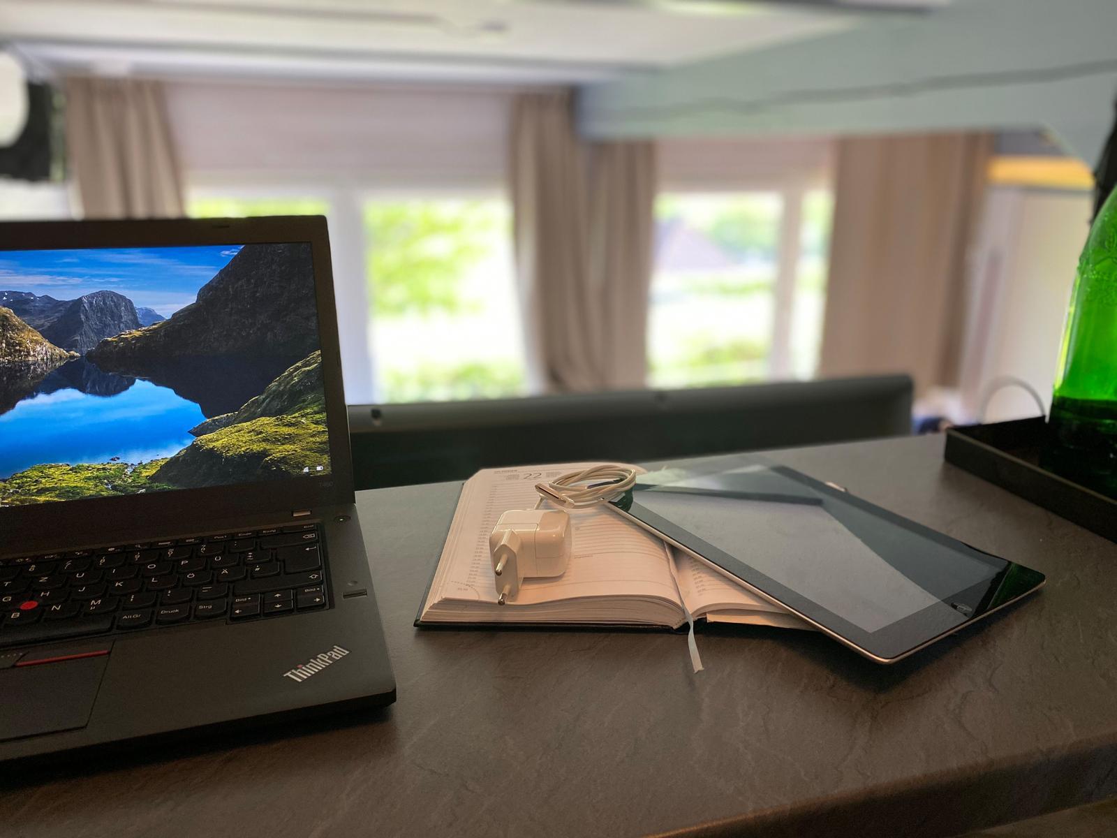 Schreibtisch mit Laptop, Notizbuch und Tablet. Fenster mit Blick auf grüne Landschaft.