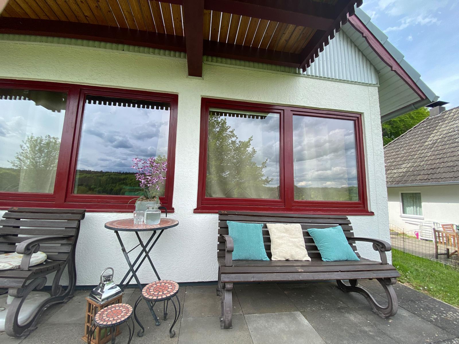 Terrasse mit Holzbank, Tisch und Stühlen. Rote Fensterrahmen und grüne Landschaft im Hintergrund.