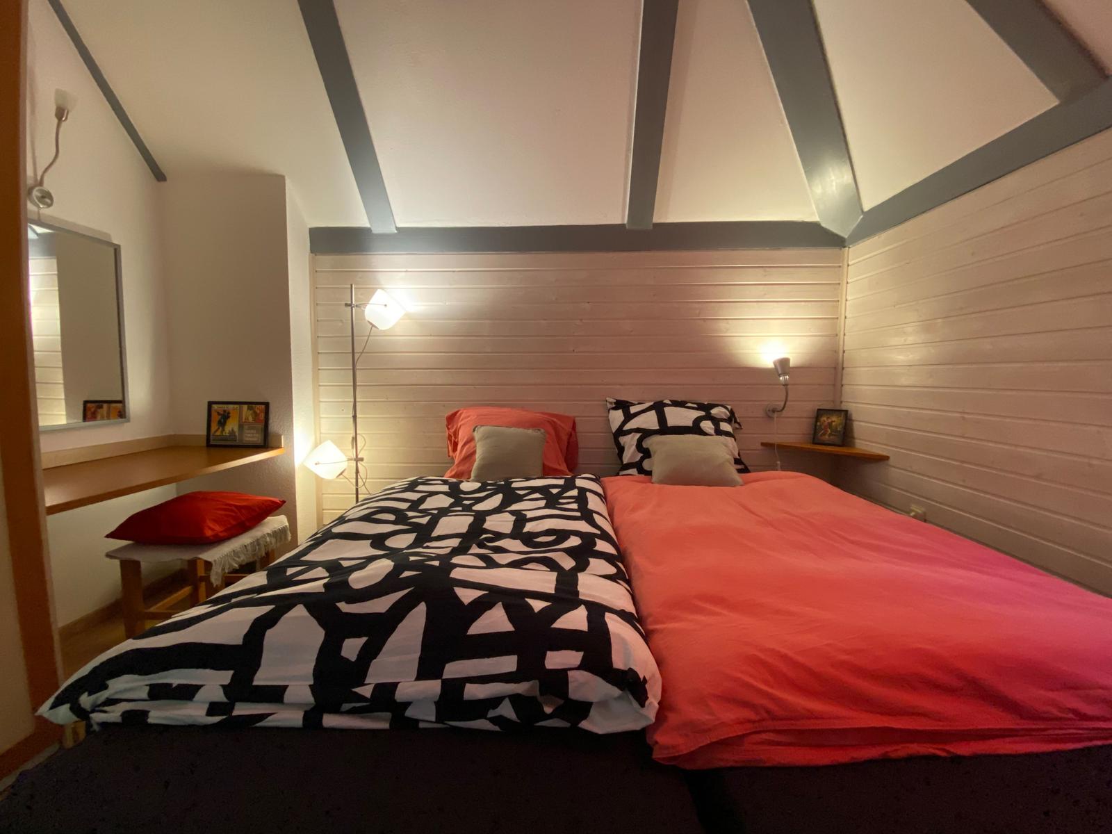 Schlafzimmer mit zwei Betten, Holzwänden und Deckenbalken. Beleuchtung und Spiegel sind vorhanden.
