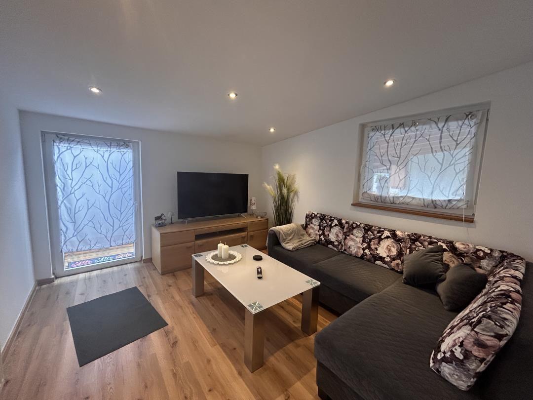 Wohnzimmer mit Sofa, TV und Holzboden. Fenster mit Blattvorhängen.