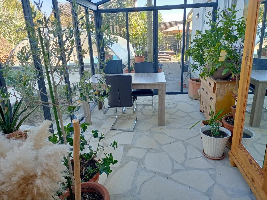 Glasveranda mit Tisch, Stühlen und Pflanzen. Steinboden und Blick auf Garten.