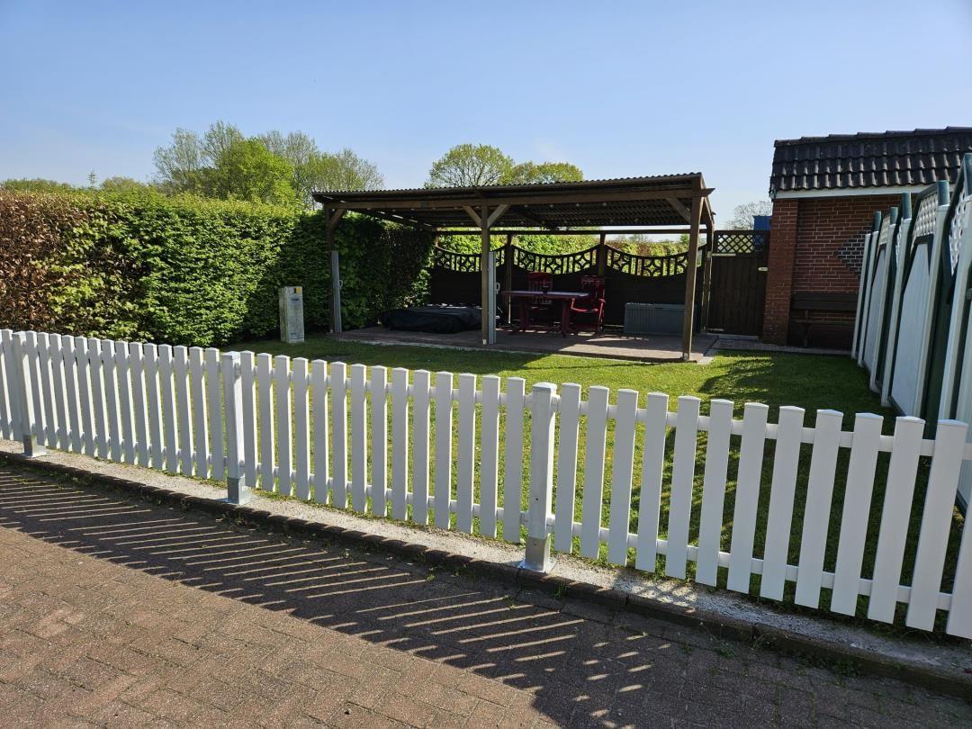 Hinterhof mit weißen Zaun, Pavillon und Gartenmöbeln. Grünflächen und Hecke sichtbar.
