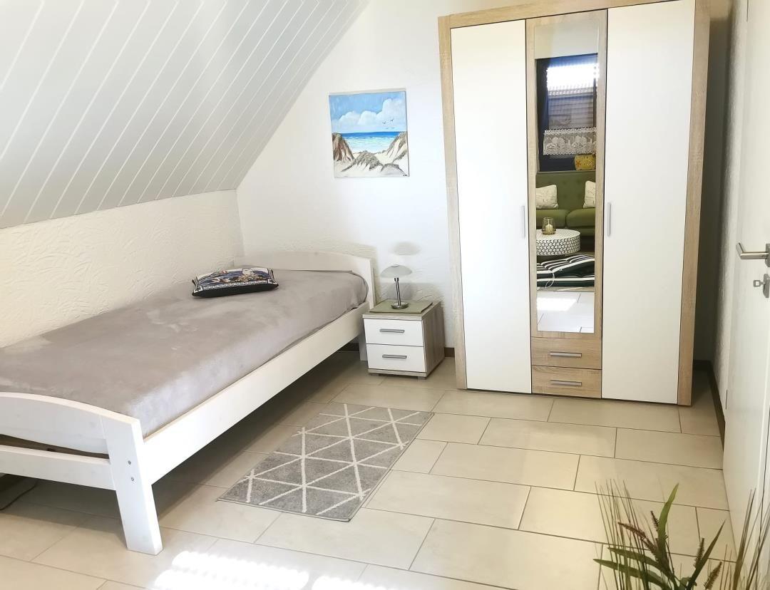 Schlafzimmer mit Einzelbett, Kleiderschrank und Teppich auf Fliesenboden.