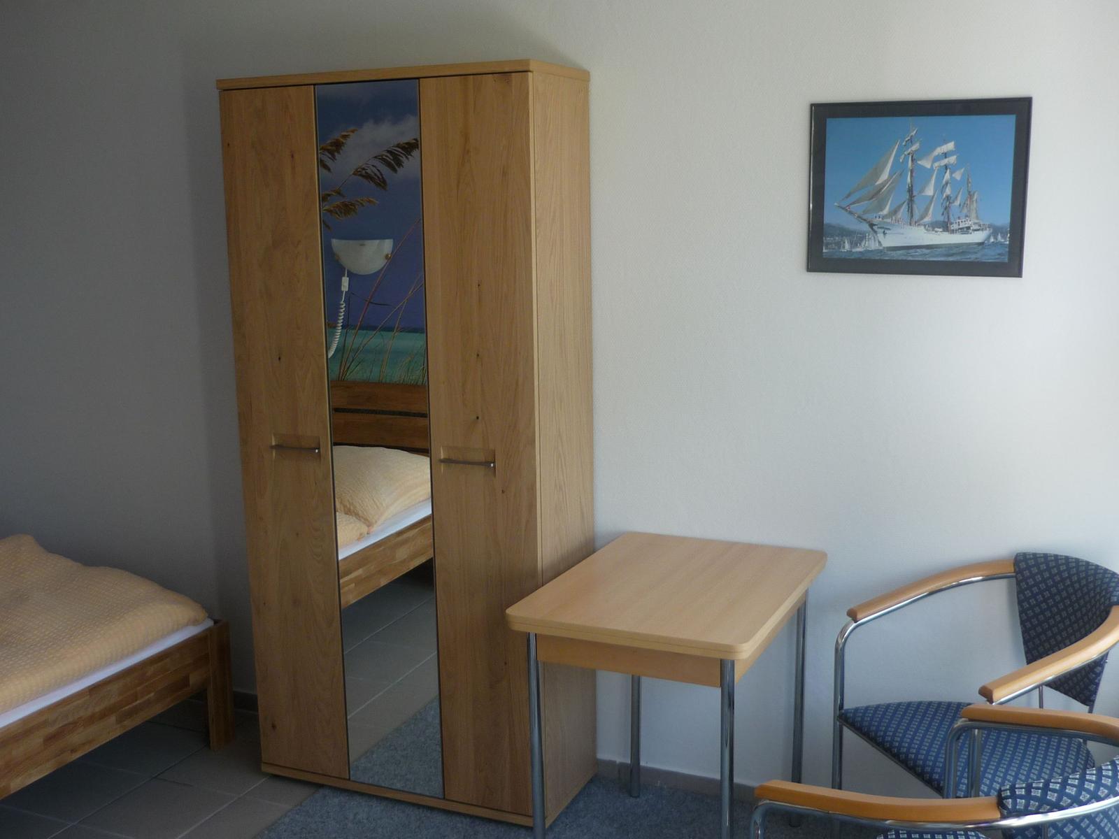 Schlafzimmer mit Bett, Kleiderschrank, Tisch und Stuhl. Wandbild zeigt Segelschiff.