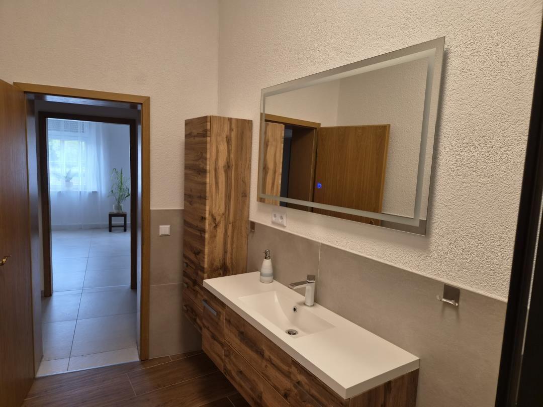 Badezimmer mit Waschbecken, Spiegel und Holzmöbel. Durchgang zu Wohnbereich.