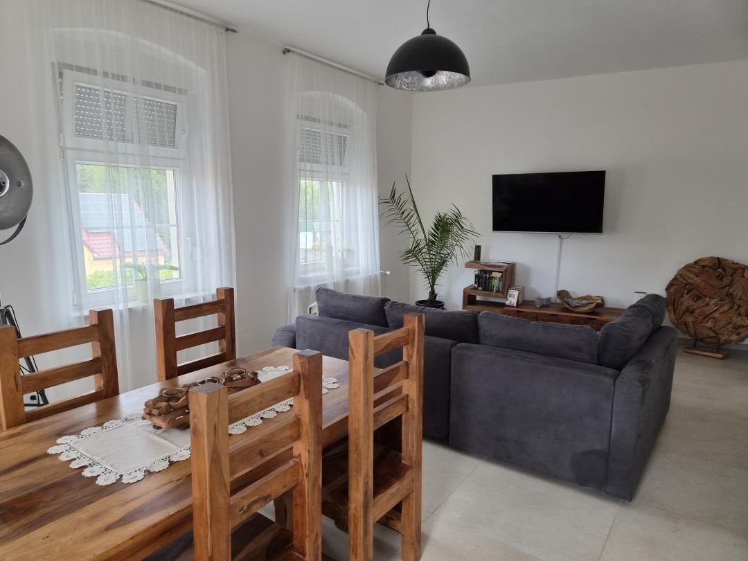 Wohn- und Essbereich mit Holztisch, Sofa und Fernseher. Fenster mit weißen Vorhängen.