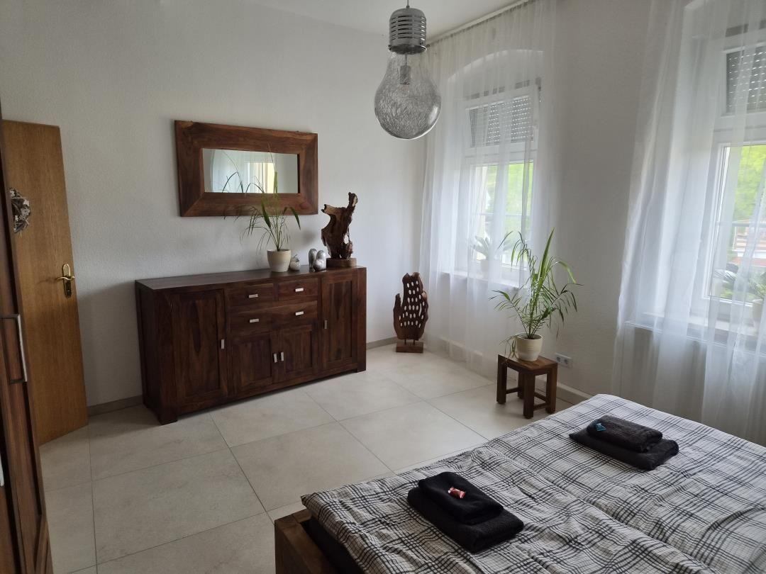 Schlafzimmer mit Bett, Holzschrank, Spiegel und Pflanzen. Fenster mit weißen Vorhängen.