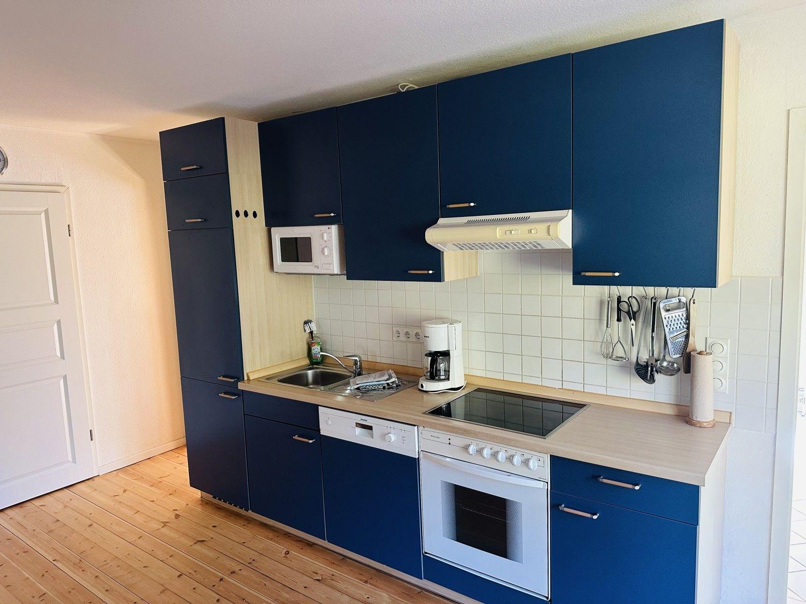 Küche mit blauen Schränken, Edelstahlspüle und Kochfeld. Holzboden und weiße Fliesen.