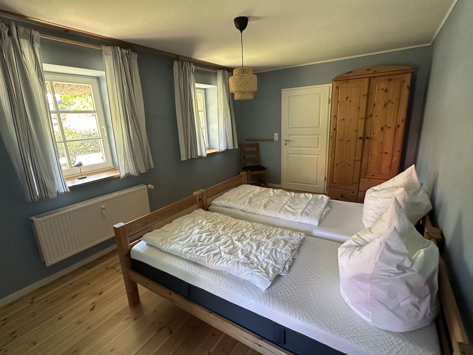 Zimmer mit zwei Betten, Holzboden und Fenstern. Holzschrank und Deckenleuchte.