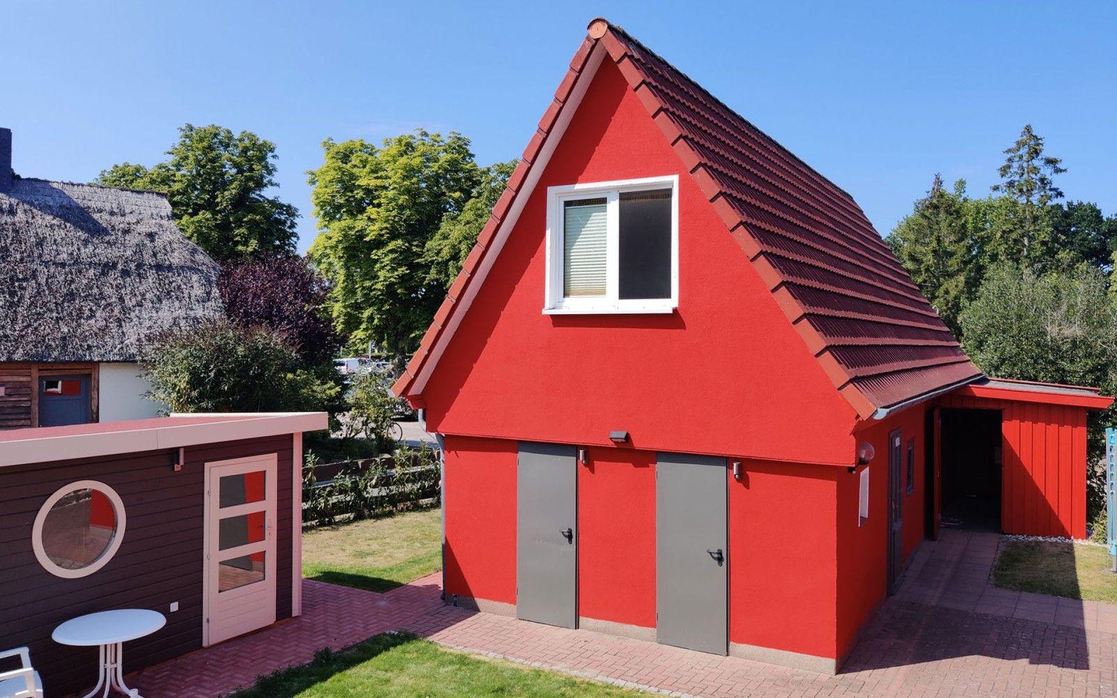 Rotes Haus mit Dach, Fenster und zwei Türen. Neben einem Gartenhaus mit runder Fensteröffnung.