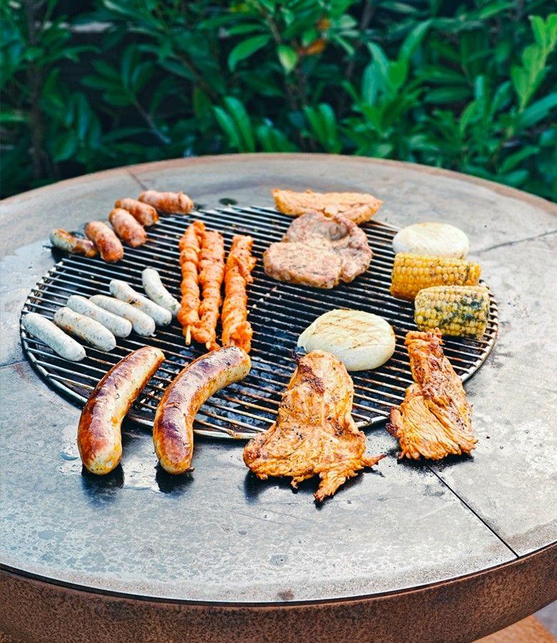 Rundes Grillgitter mit Würsten, Fleisch und Mais auf Steinplatte im Garten