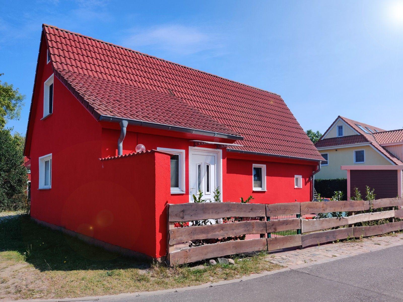 Rotes Haus mit Dach und Holzzaun. Hintergrund: weiteres Haus und blauer Himmel.