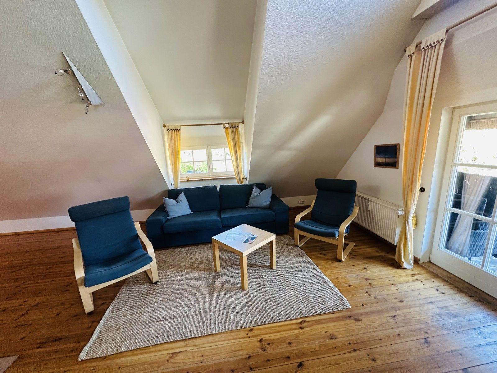 Wohnzimmer mit Sofa, zwei Sesseln, Tisch und Holzboden unter schrägen Wänden.