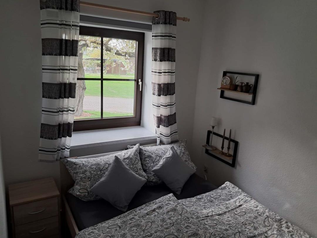 Schlafzimmer mit Bett, Fenster und Vorhängen. Außen sichtbar Grünfläche und Baum.