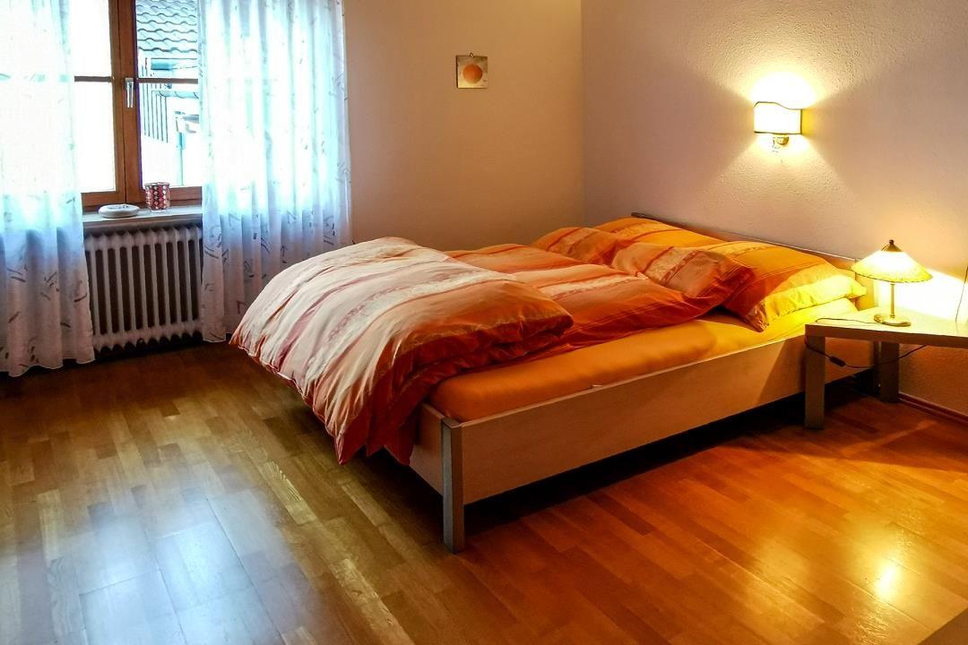 Schlafzimmer mit Bett, Nachttisch, Lampe und Fenster mit Vorhängen.