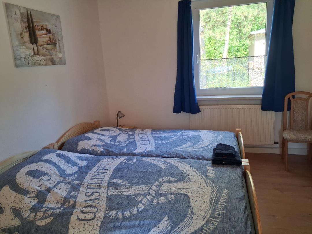 Zwei Einzelbetten mit blau-weißem Bettbezug, Fenster mit blauen Vorhängen und Stuhl.