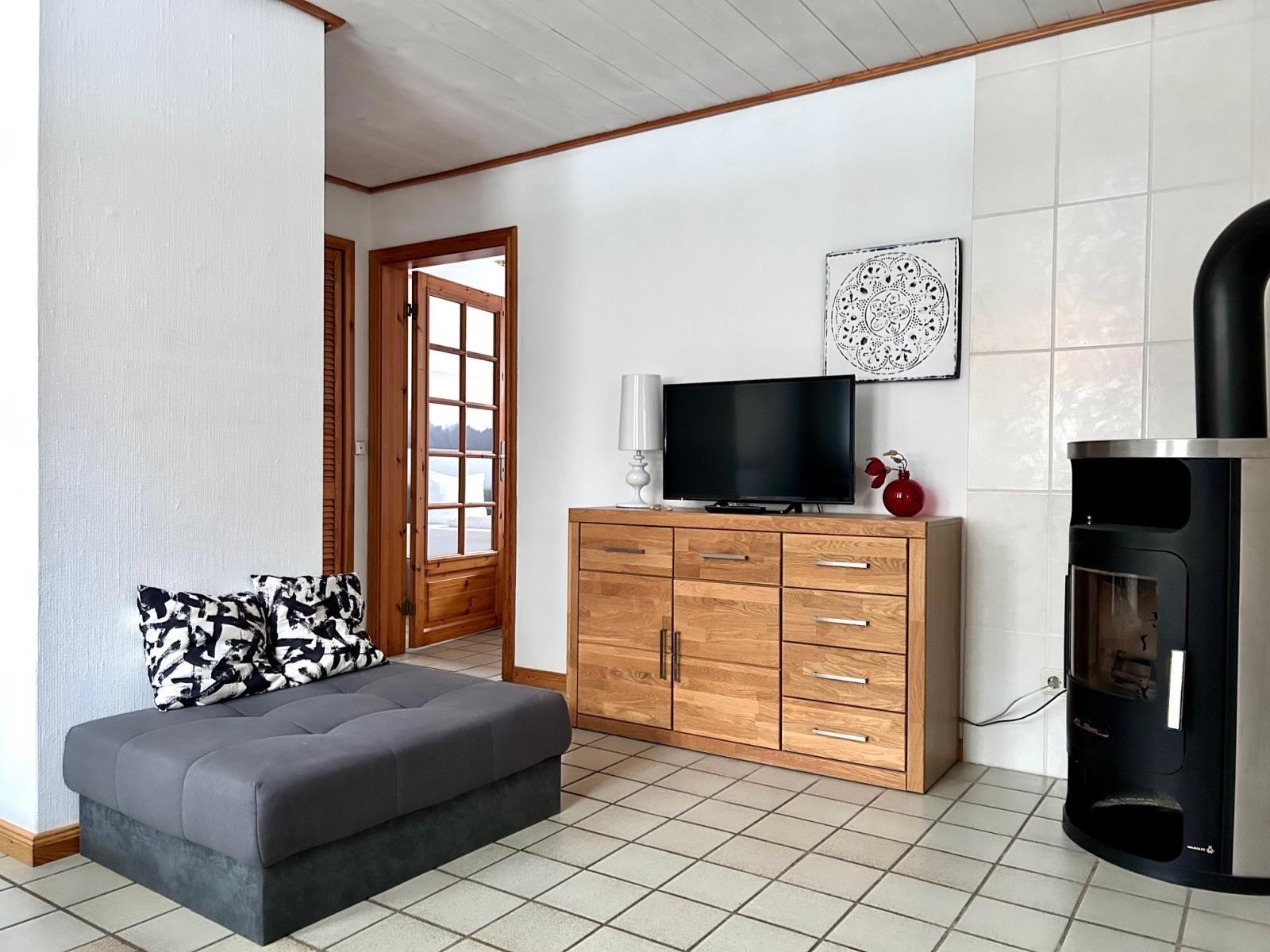 Ein Wohnzimmer mit grafer Sofa, Holzschrank, Fernseher und Kaminofen.