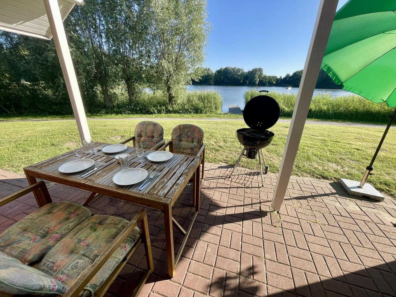 Patio with table, chairs, grill, and green umbrella by the lake.