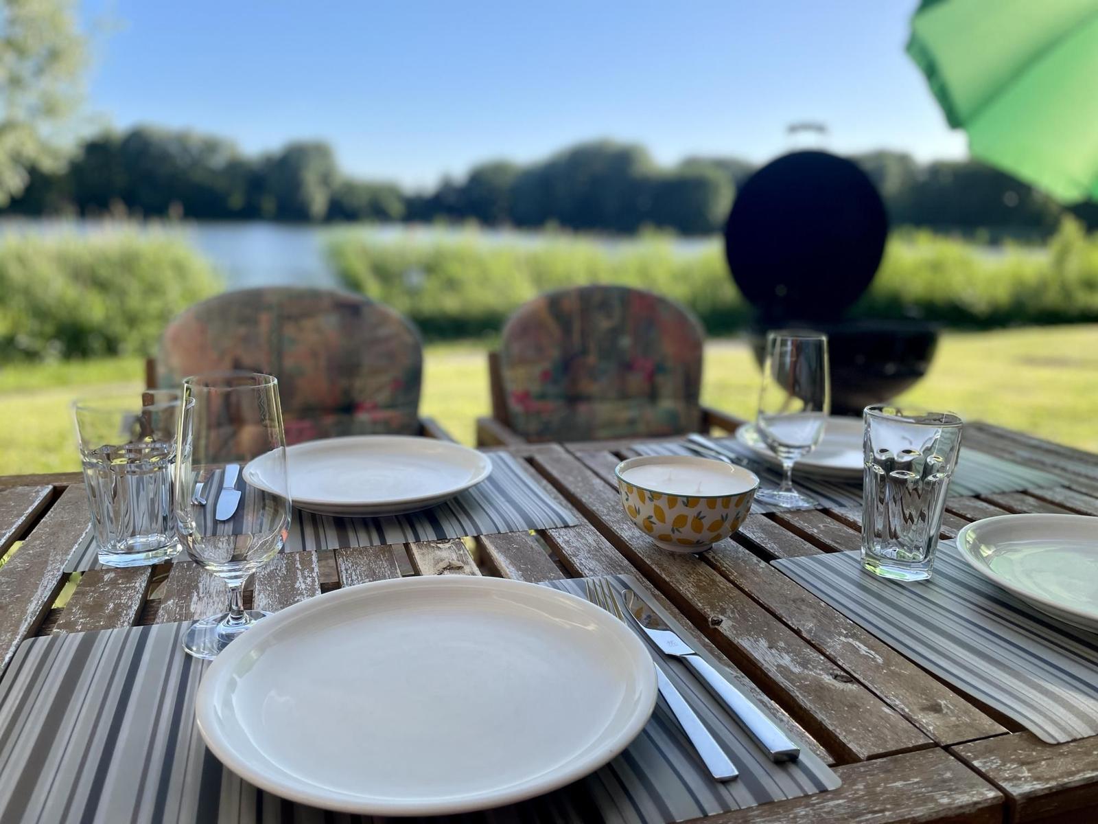 Set table for four on wooden deck by lake. Grill and green umbrella visible.