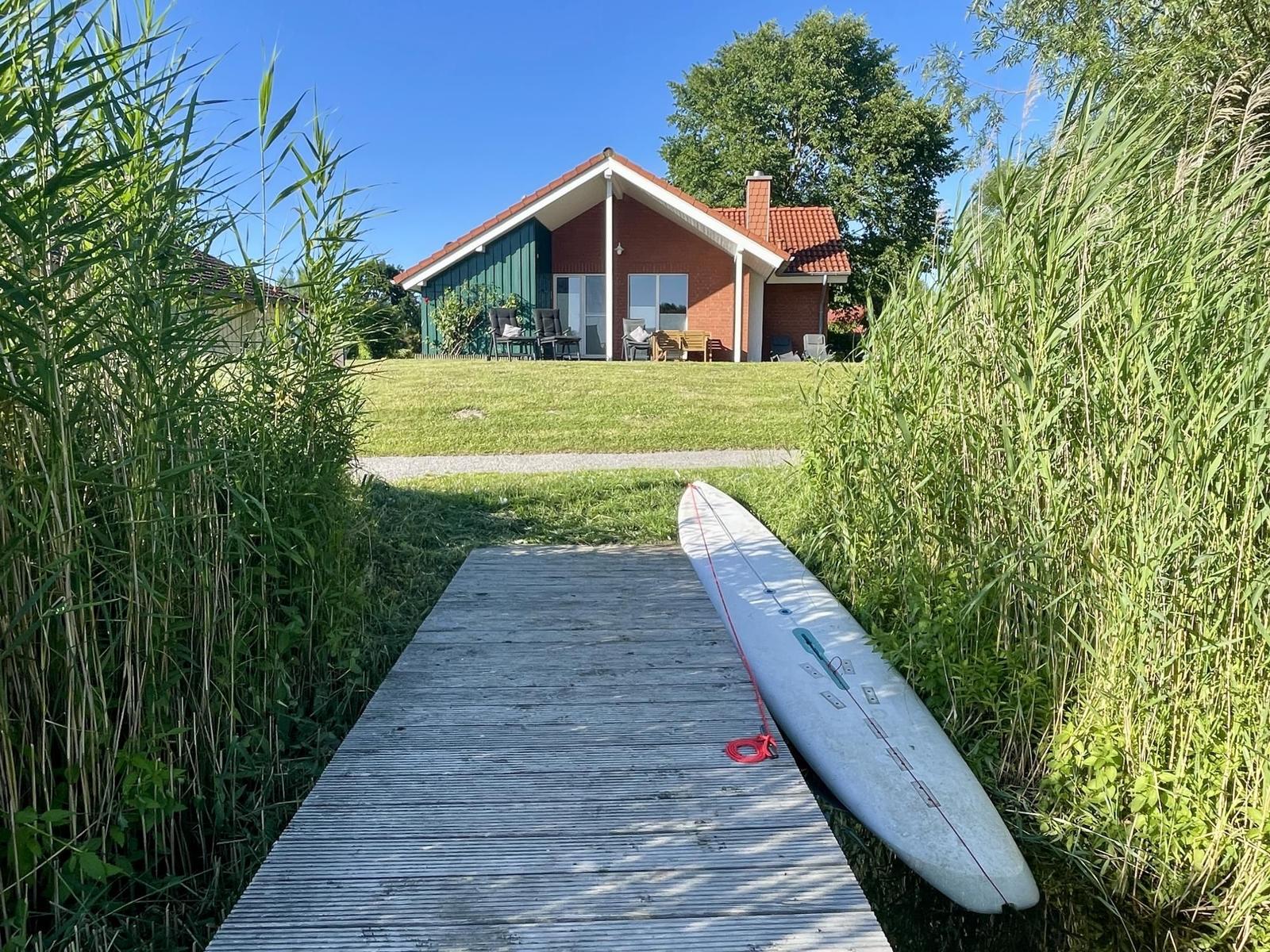 A surfboard rests on a wooden path leading to a house with a garden.