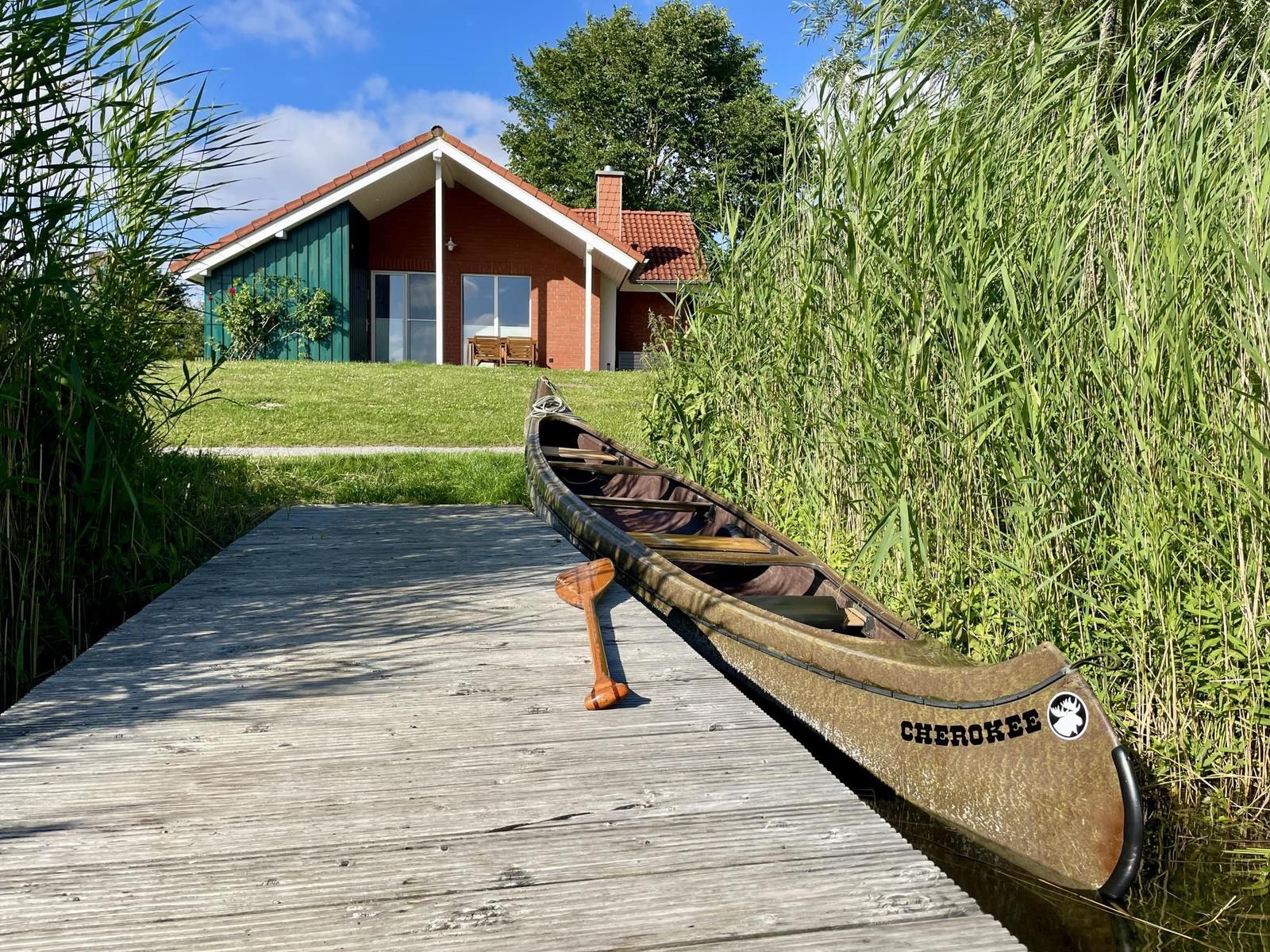 A canoe sits on a wooden dock in front of a house with a red roof.