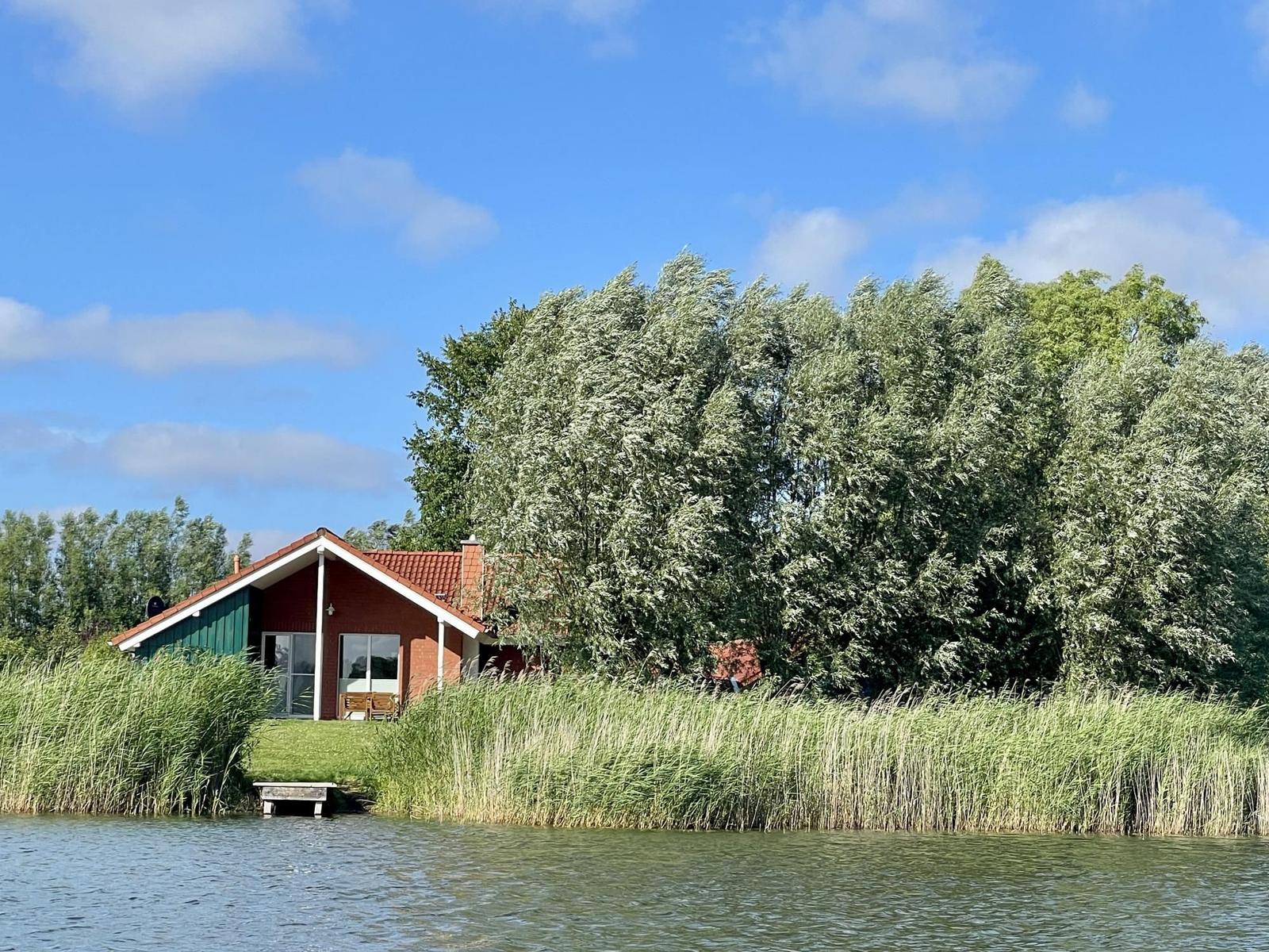 A house by the water with trees and tall grass in the foreground.