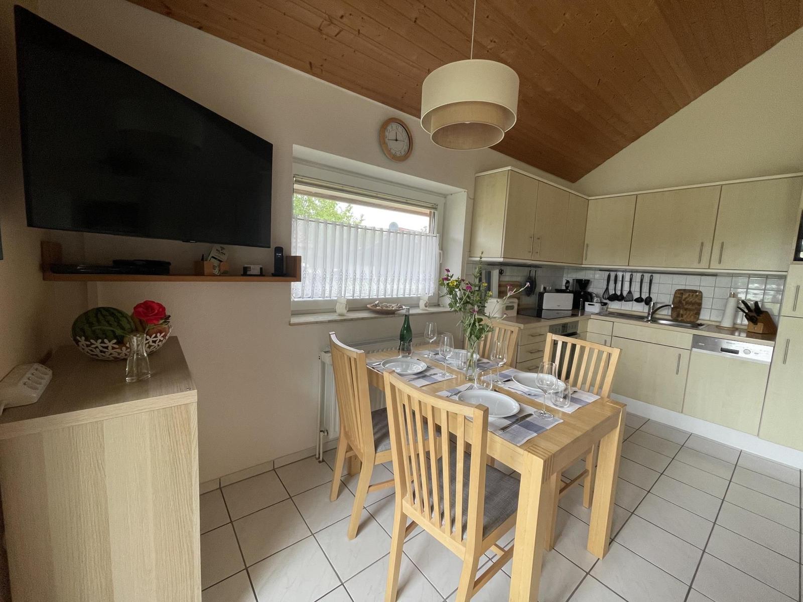Dining area with table, chairs, and kitchen. TV, window, and wooden ceiling are visible.