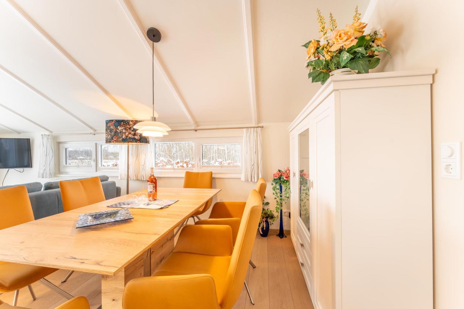 Dining area with wooden table, orange chairs, and windows overlooking the landscape.