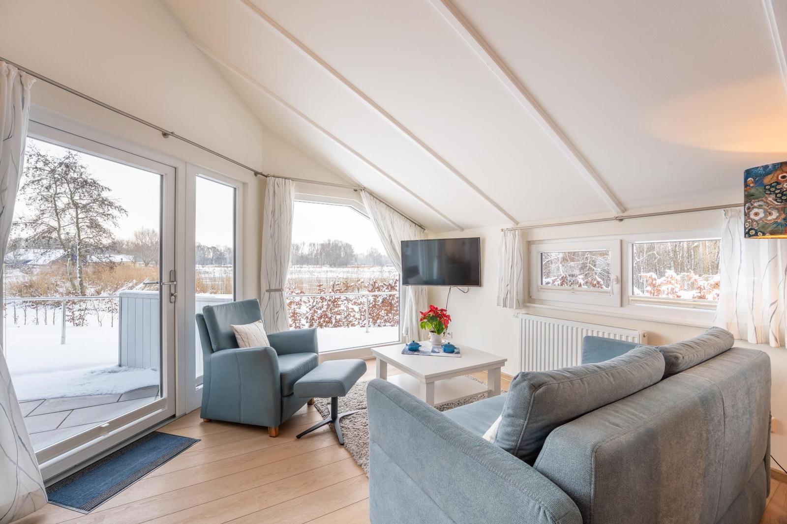 Living room with sofa, armchair, and TV. View of snowy balcony and forest.