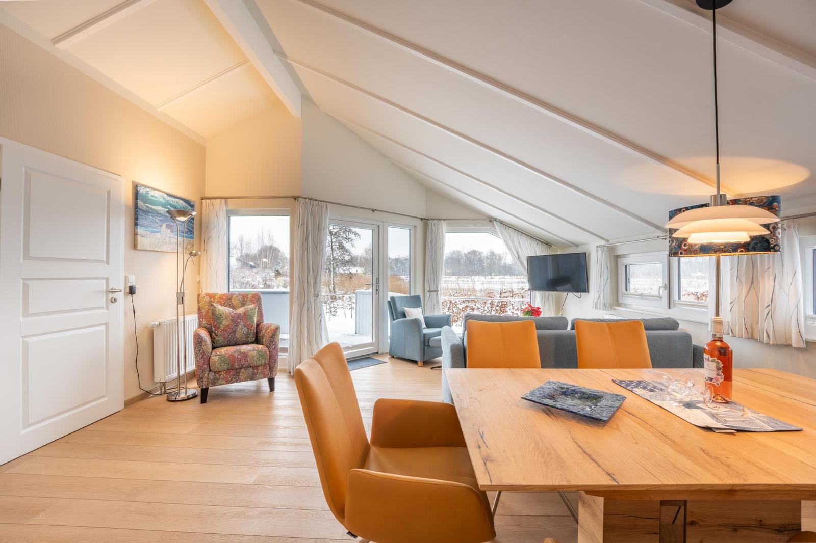 Living and dining area with wooden table, chairs, and TV. View of balcony and landscape.