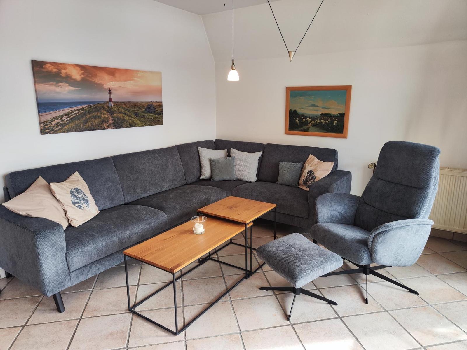 Ein Wohnzimmer mit grauem Sofa, Sitzhocker und Holztisch. An den Wänden hängen Bilder.