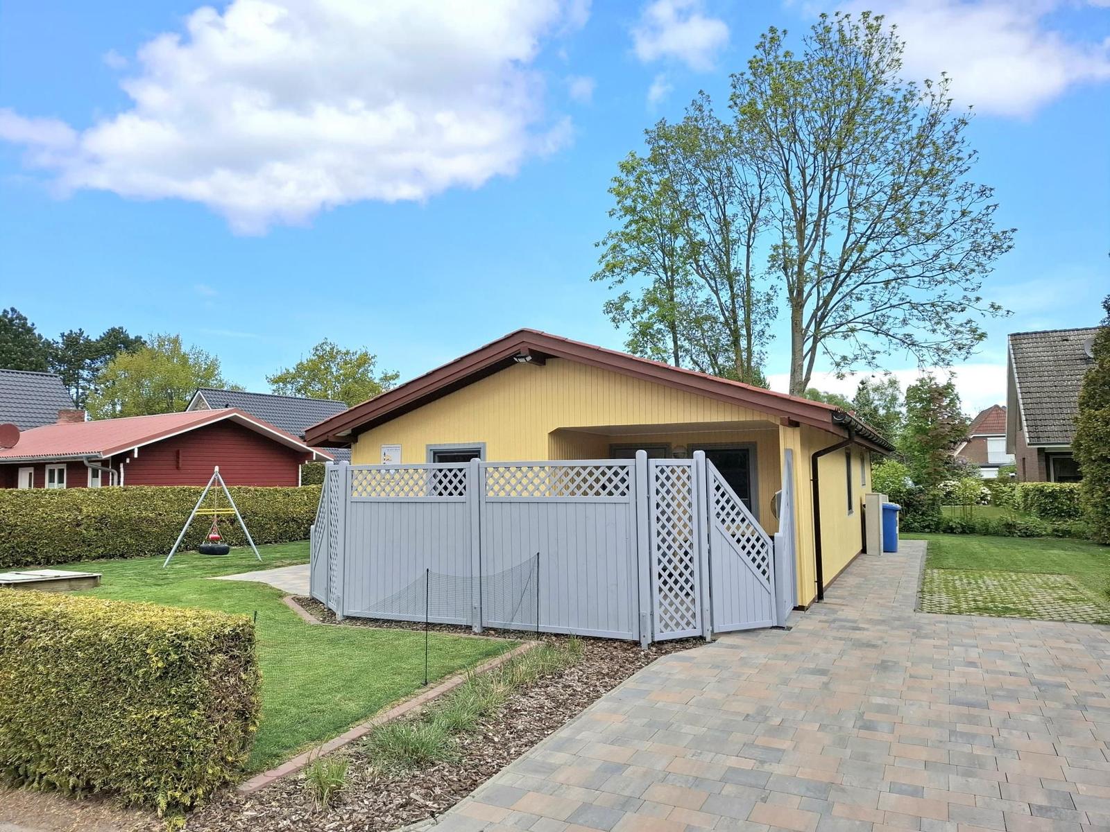 Gelbes Haus mit Gartenzaun und Spielplatz. Pflasterweg führt zur Eingangstür.