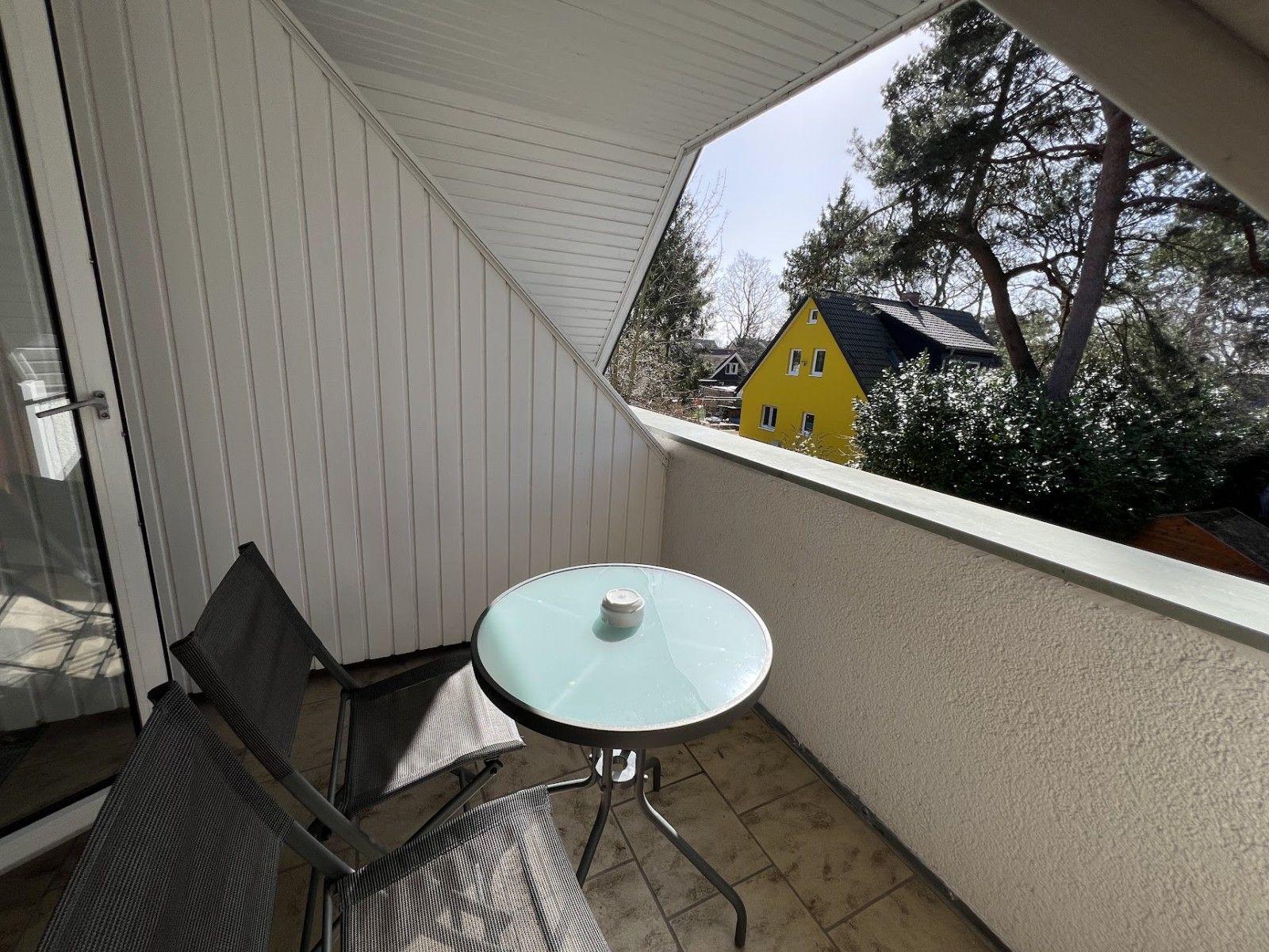 Terrasse mit Tisch und zwei Stühlen. Blick auf gelbes Haus und Bäume.